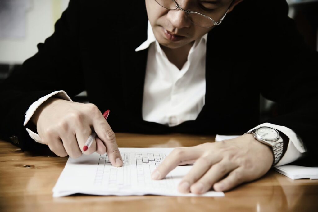 A person in a suit is writing on a sheet of paper with a red pen, focused on the document on a wooden table.