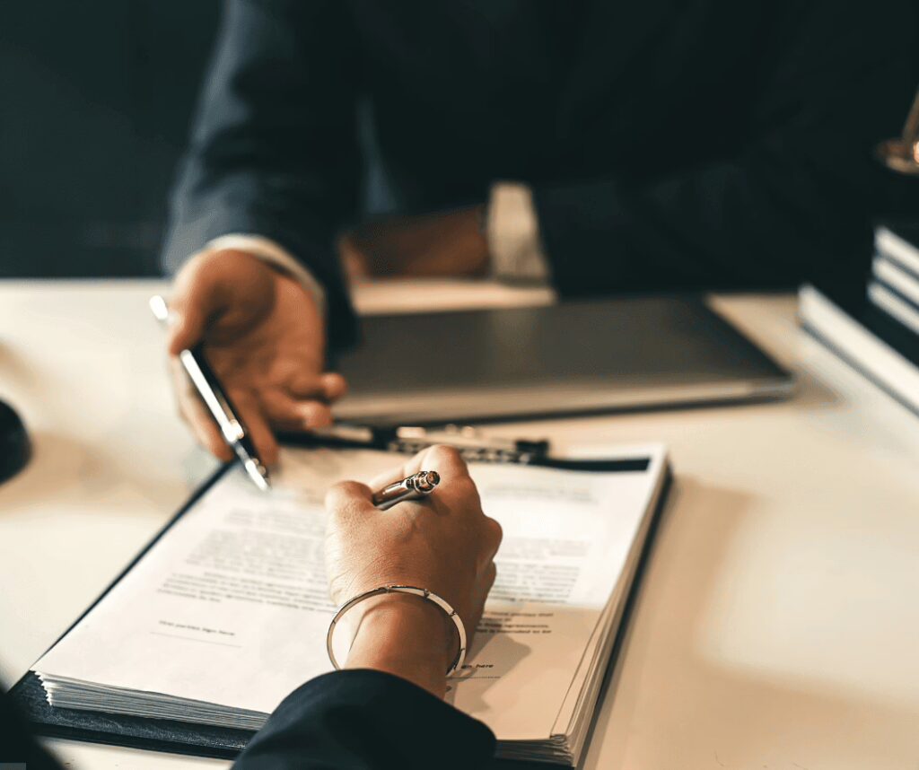 Hands of two individuals in business attire discussing a contract on a desk, one holding a pen above printed papers.