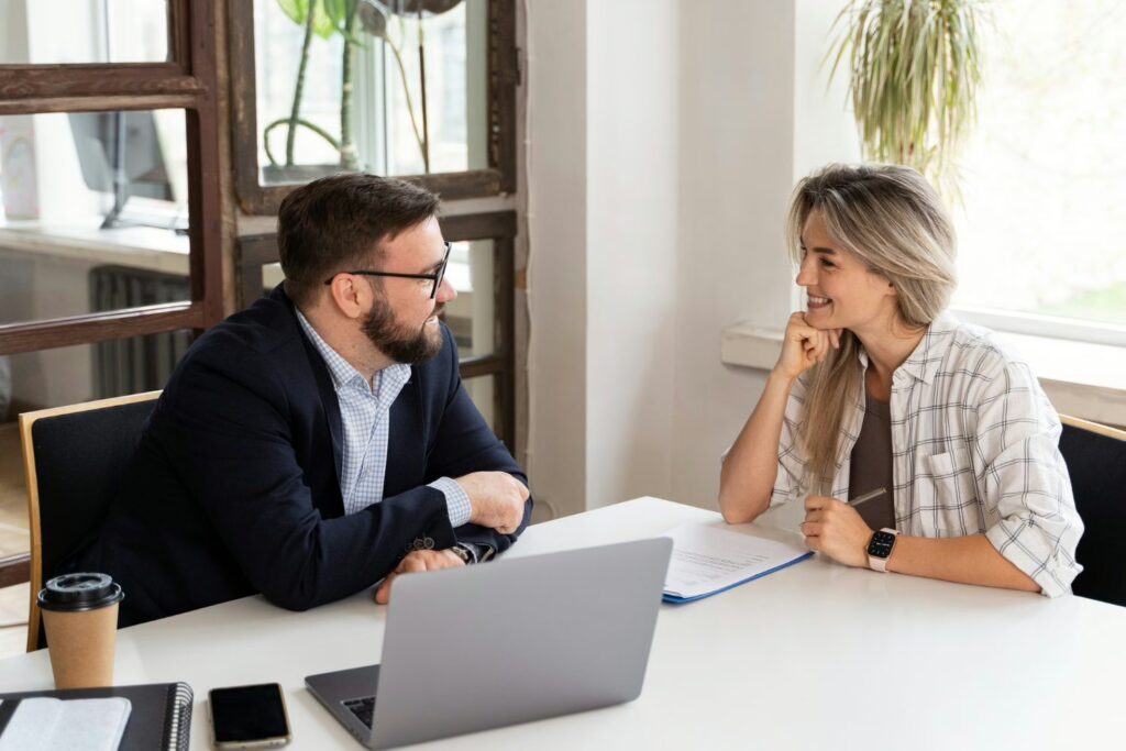Two professionals in a bright office engage in conversation, with a laptop and notes on the table between them.